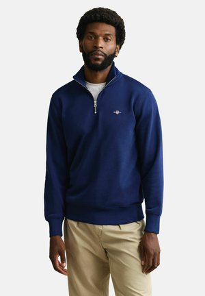 Mann mit Bart, der einen marineblauen Pullover mit Reißverschluss bis zur Brust und beige Hosen trägt, vor einem schlichten weißen Hintergrund stehend.