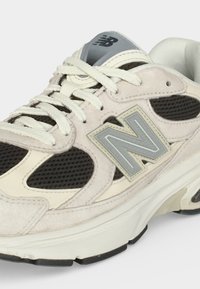 Ανοιχτό μπεζ και μαύρο αθλητικό παπούτσι New Balance με γκρι λογότυπο, πάνελ από δίχτυ, παχιά σόλα και λευκά κορδόνια.