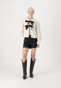 Κρεμ πλεκτό cardigan με μαύρη γραβάτα στον λαιμό, συνδυασμένο με μαύρα δερμάτινα σορτς και καφετί καουμπόικες μπότες. Λείος υφή, κοντό μήκος.