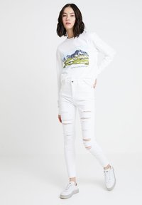 Biała bluza z grafiką góry i napisem, w połączeniu z białymi, przetartymi rurkami i białymi trampkami z szarymi akcentami.