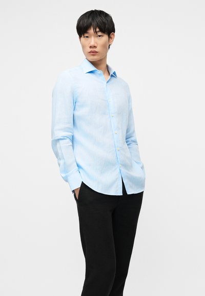 Νεαρός άνδρας που στέκεται με τα χέρια στις τσέπες, φοράει ένα γαλάζιο πουκάμισο με κουμπιά και μαύρο παντελόνι, με φόντο ένα λευκό τοίχο.