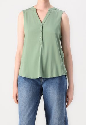 Blouse sans manches vert clair avec un col en V et trois boutons, portée avec un jean en denim bleu sur une personne debout.