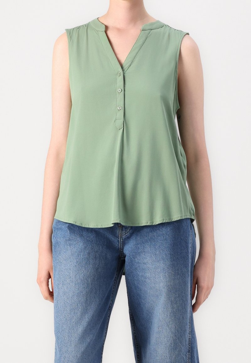 Blouse sans manches vert clair avec un col en V et trois boutons, portée avec un jean en denim bleu sur une personne debout.