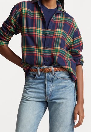 Person iført en blå, rød, grøn og gul ternet skjorte med knapper over en mørkeblå top, lyseblå jeans og et flettet brunt bælte.