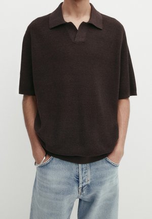 Homme portant un polo en maille marron foncé à manches courtes avec col, jean bleu clair délavé, et un bracelet argenté, les mains dans les poches.