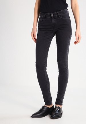 Henkilö pukeutunut mustiin kapeisiin farkkuihin, joissa on edessä ja takana taskut, yhdistettynä mustiin nahkaisiin liukusukkiin, joissa on vetoketjut.