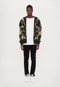 Felpa nera con zip e motivo floreale giallo, t-shirt bianca, pantaloni neri e sneakers bianche. Tessuto morbido, vestibilità casual.
