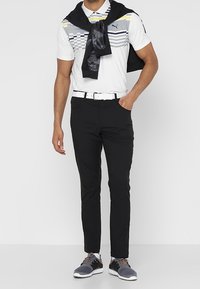 Polo blanc avec un motif à rayures grises et jaunes, pantalon noir, et baskets grises. Une veste noire drapée sur les épaules complète le look.