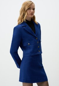 Blue textured tweed jacket with a cropped design, two front pockets, and six buttons, paired with a matching skirt.