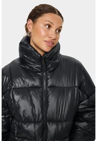 Melnā pūšamā jaka ar augstu apkakli, spīdīgu tekstūru, rāvējslēdzēja aizdari un šūtu dizainu ar polsterētām sadaļām siltuma nodrošināšanai.