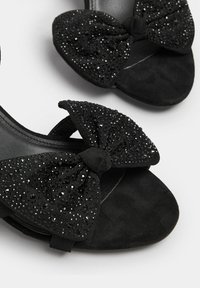 Black heeled sandals featuring a large bow with shiny rhinestones, soft suede-like material, open toe design, and flat heel.