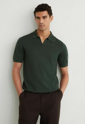 Homme aux cheveux bouclés et foncés portant un polo vert foncé à manches courtes et un pantalon foncé, debout les mains dans les poches devant un mur blanc.