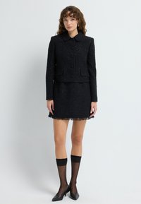 Mustat tweed-takit, joissa on kaulukset, nappikiinnitys ja kaksi taskua, yhdistettynä fringettömään mustaan hameeseen ja läpinäkyviin mustiin polvisukkiin.