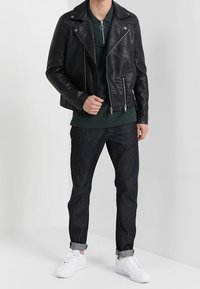 Černá kožená motorkářská bunda se stříbrnými zipy, vrstvená přes tmavě zelený top, v kombinaci s tmavými džínami a bílými teniskami.