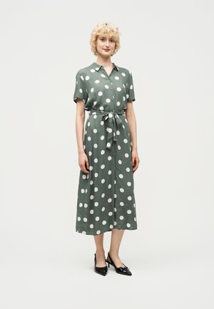 Γυναίκα με κοντά, σγουρά ξανθά μαλλιά που φοράει πράσινο μίντι φόρεμα με λευκές πουά, δεμένο στη μέση, και μαύρα μυτερά ψηλοτάκουνα.