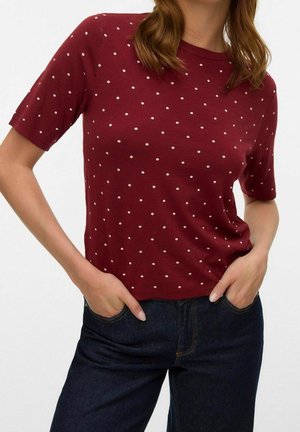 Kobieta ubrana w bordową bluzkę z krótkim rękawem w białe kropki oraz ciemnoniebieskie dżinsy, z rękami wsuniętymi do przednich kieszeni.