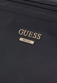 Tumed kangast kott, millel on kuldne "GUESS" logo ja "EST.1981" silt, sujuva tekstuuriga ja tõmblukuga sulgur.