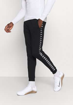 Homme portant un pantalon noir fuselé avec une bande blanche avec logo sur les côtés, une chemise blanche à manches longues, des chaussettes blanches et des chaussures de sport blanches.