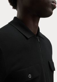 Sort strikket polo-shirt med lynlås og to brystlommer med knapdetaljer. Materialet har en tekstureret overflade.