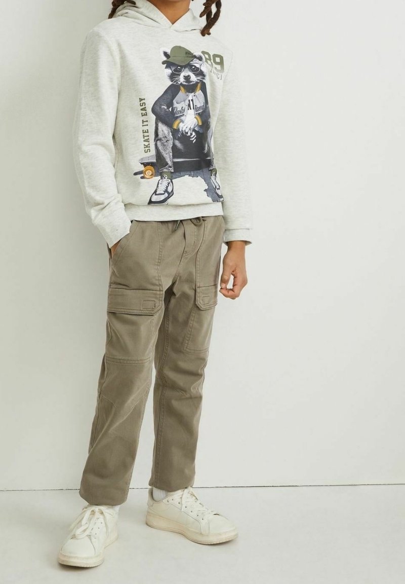 Enfant portant un sweat à capuche gris clair avec un motif de raton laveur en train de faire du skateboard, un pantalon cargo kaki et des baskets blanches, debout contre un mur blanc.