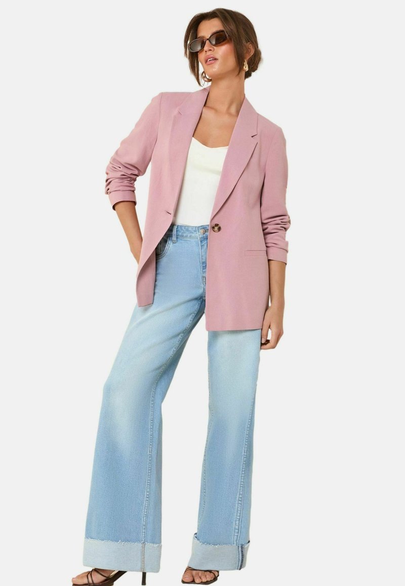 Svetlo roza blazer s enim gumbom, nosi se preko belega topa in svetlo modrih širokih hlač, ki imajo strgane manšete in sproščen kroj.