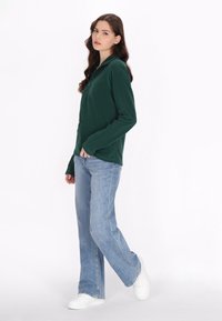 Maglia verde a maniche lunghe con colletto, abbinata a jeans dritti azzurri chiari e sneakers bianche, su uno sfondo semplice.
