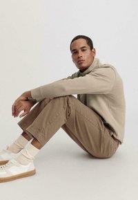 Felpa beige con colletto alto, pantaloni beige e sneaker bianchi. L'outfit presenta una vestibilità rilassata e un tessuto testurizzato.