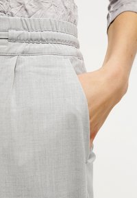 Ljust grå, vävda byxor med elastisk midja och sidofickor. Texturen är slät och visar ett subtilt mönster.