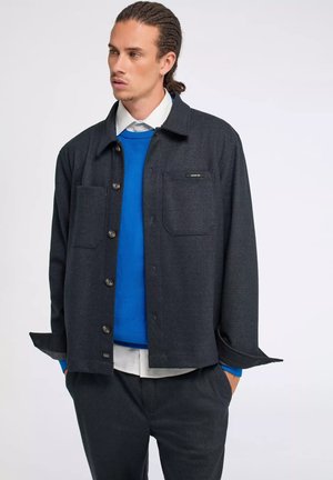 Giacca camicia grigia scura con maniche lunghe, due tasche sul petto e un'etichetta. Indossata sopra un maglione blu e abbinata a pantaloni coordinati.