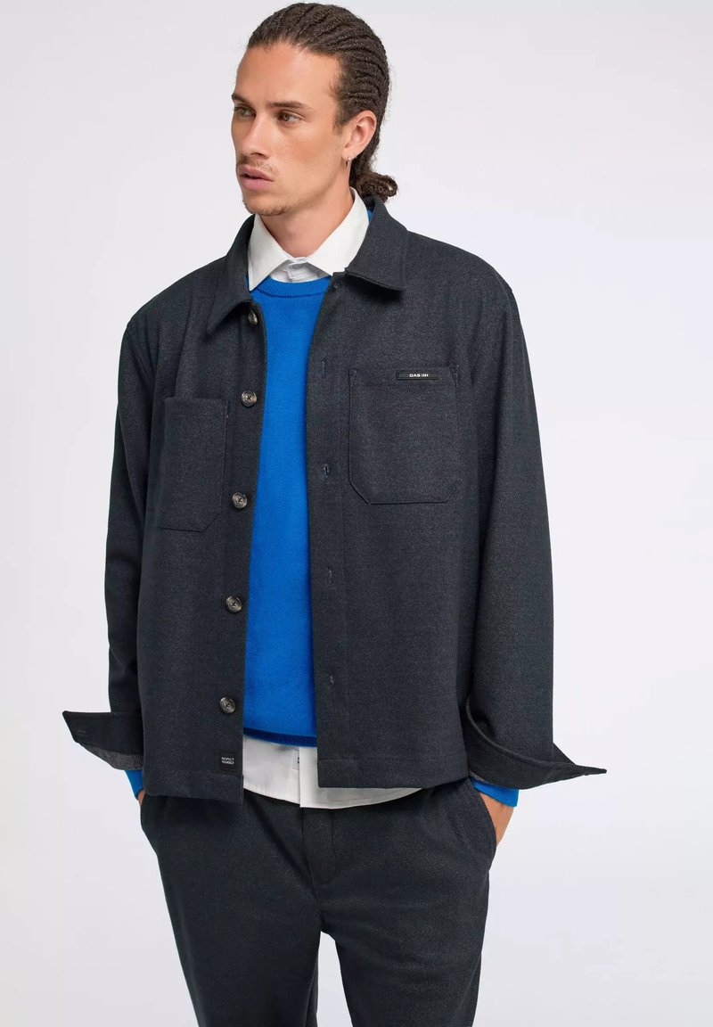 Giacca camicia grigia scura con maniche lunghe, due tasche sul petto e un'etichetta. Indossata sopra un maglione blu e abbinata a pantaloni coordinati.