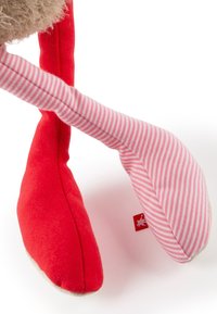 Stuffed toy with fuzzy beige body, red limbs, and one pink striped leg. Features small tag with a logo. Soft fabric texture throughout.