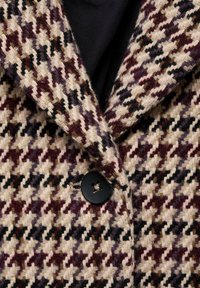 Vävd kappa med ett hundtandsmönster i kräm, burgundy och svart, med en stor svart knapp och textil med struktur.