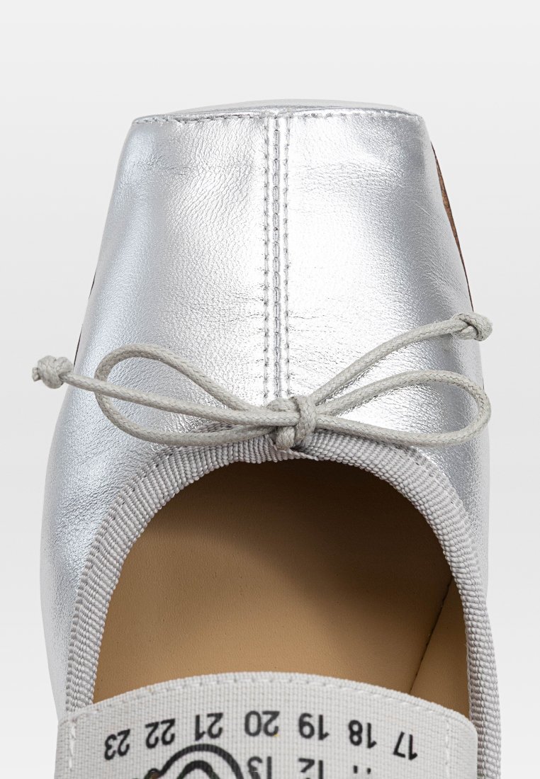 Scarpa piatta metallica argento con punta arrotondata, fascia elastica grigia testurizzata e un dettaglio a fiocco sulla parte frontale. Interno beige.
