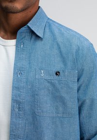 Lichtblauw chambray overhemd met een button-down kraag, een enkele borstzak en zwarte knoopdetails. Zachte textuur en een licht getailleerd ontwerp.