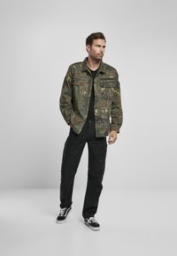 Tarnjacke mit zwei Brusttaschen, kombiniert mit schwarzen Cargo-Hosen und schwarzen Sneakern. Die Jacke hat einen knöpfbaren Schnitt und eine entspannte Passform.