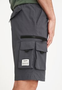 Grå cargoshorts med elastiskt midjeband, flera sidofickor och dragkedja fram. Har en liten tygetikett med text som är fäst.