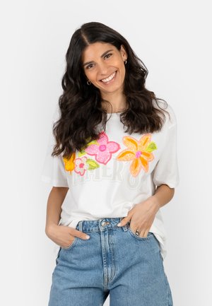 Smiling woman with long dark hair wears white T-shirt with colorful flowers and blue jeans, hands in pockets against plain background.