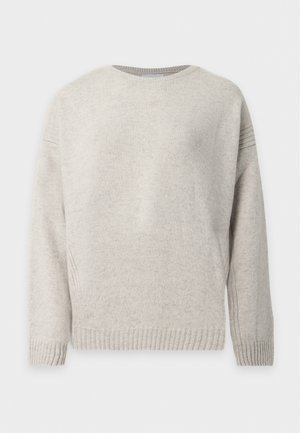 Frau steht, trägt einen beigen Pullover, weite cremefarbene Hosen, braune spitze Schuhe und hält eine kleine braune Lederschultertasche.