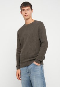 Suéter de color verde oliva oscuro texturizado con cuello redondo y puños acanalados, usado con jeans de color azul claro, de pie contra un fondo blanco.