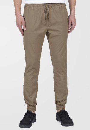 Homme portant un pantalon de jogging beige avec une taille à cordon et des poignets élastiques, associé à des baskets noires sur un fond uni.