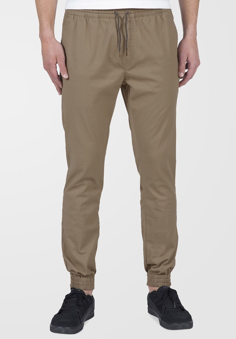 Homme portant un pantalon de jogging beige avec une taille à cordon et des poignets élastiques, associé à des baskets noires sur un fond uni.