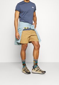 Béžové kraťasy, tmavě modré tričko s kulatým výstřihem a logem, světle modrá mikina uvázaná v pase, vzorované ponožky a béžové sportovní boty.