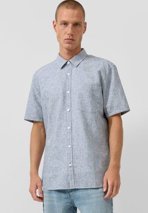 Mężczyzna w jasnoniebieskiej koszuli na krótki rękaw z guzikami i jasnych dżinsach, stojący na tle jednolitego szarego tła.