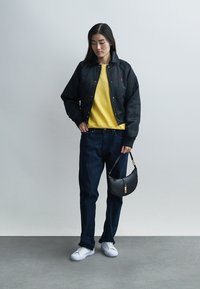 Veste légère noire, pull côtelé jaune, jean foncé, baskets blanches et petit sac à main noir. Textures lisses, coupe décontractée.