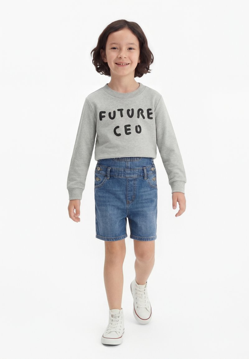 Enfant aux cheveux foncés mi-longs portant un sweat-shirt gris "FUTURE CEO", un short en jean et des baskets blanches, marchant vers l'avant.
