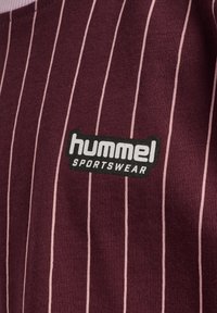Burgundy sportkläder med vertikala rosa ränder; har en svart rektangulär logotyp med vit text "hummel SPORTSWEAR." Mjuk textur.