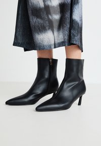 Črne usnjene gležnjarje s koničastim nosom in zadrgo ob strani, zaključene s koničasto peto. Gladka tekstura in eleganten dizajn.