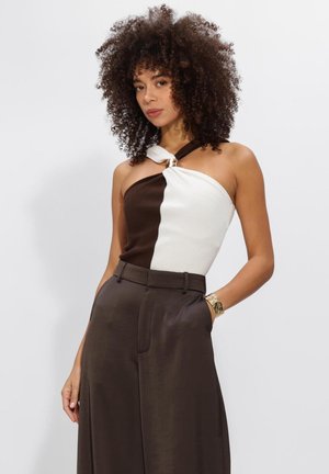 Vrouw met krullend haar draagt een mouwloze, tweekleurige top in bruin en wit met een broek met hoge taille in bruin en een gouden armband, poserend tegen een effen achtergrond.