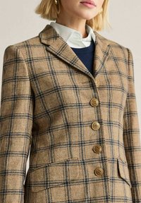 Nainen, jolla on yllään beige ruudullinen bleiseri tummilla viivoilla, ruskeat napit ja valkoinen kauluspaita alla.
