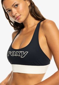 Čierny športový top s bielym prúžkom, ktorý na prednej strane obsahuje výrazné logo "roxy". Vyrobený z hladkého, elastického materiálu, navrhnutý pre pohodlie.